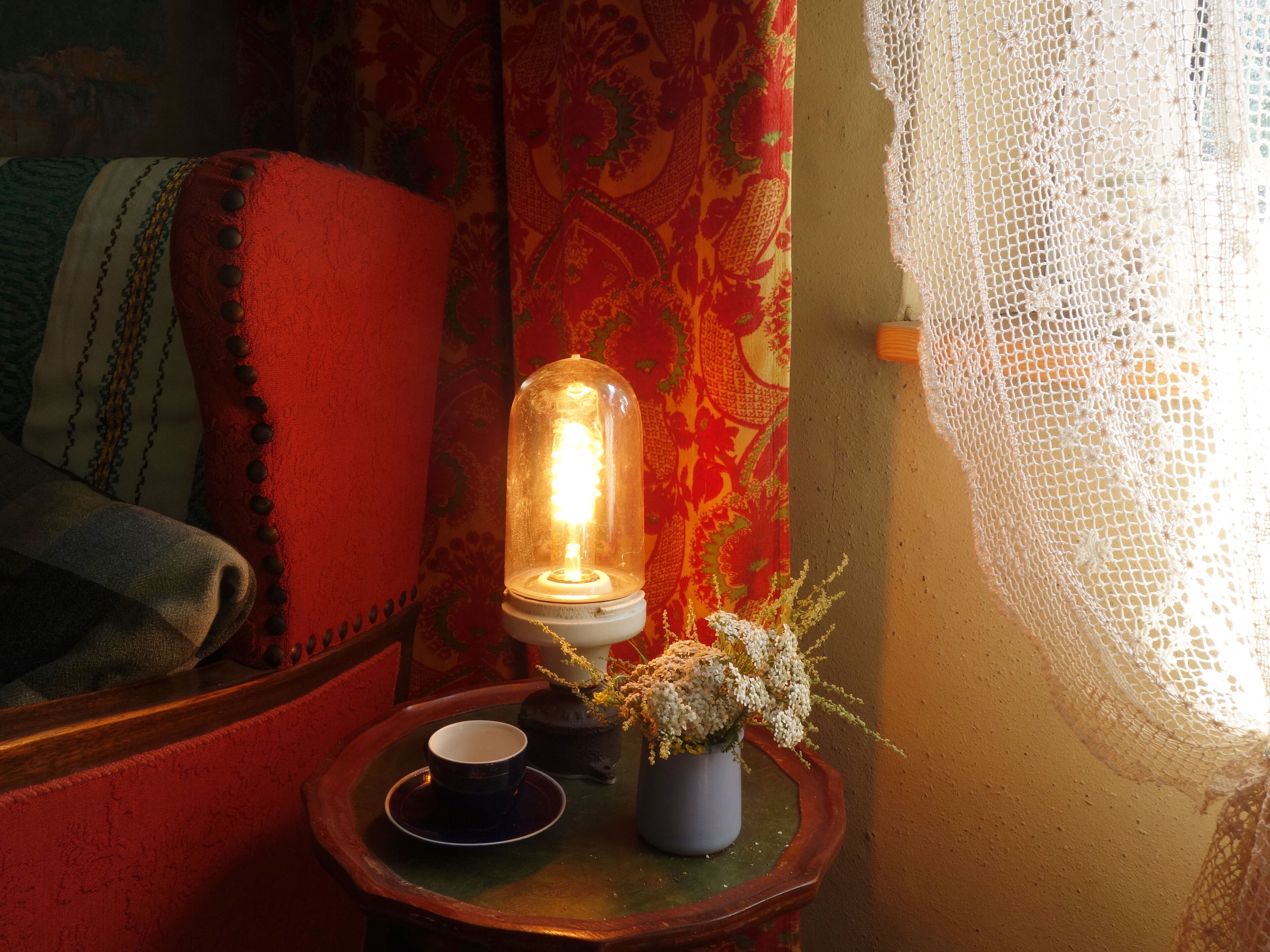 A cozy room with a vintage lamp, a small table with a cup and flowers, and patterned curtains.