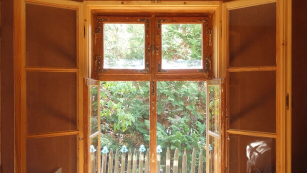 Une pièce dotée d’une grande fenêtre à cadre en bois, d’un banc en bois et offrant une vue sur une clôture en bois et la verdure environnante.