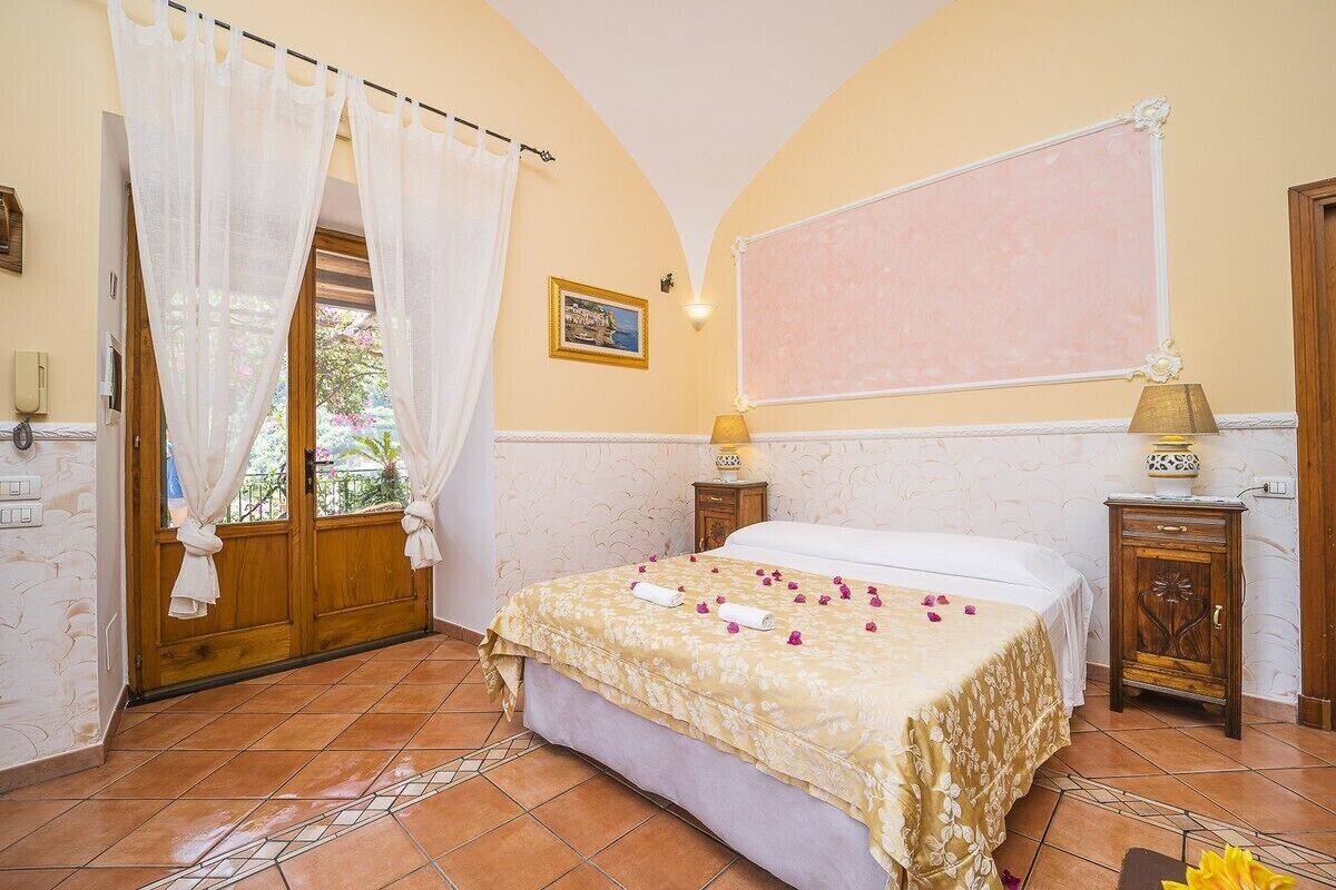 A bedroom with a bed, a window with curtains, a bedside table with a lamp, and a wall-mounted light.