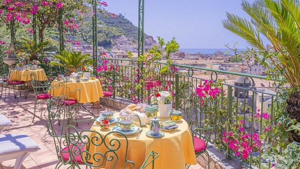 Una terrazza all'aperto con tovaglie gialle, sedie in metallo e vista su una cittadina costiera.