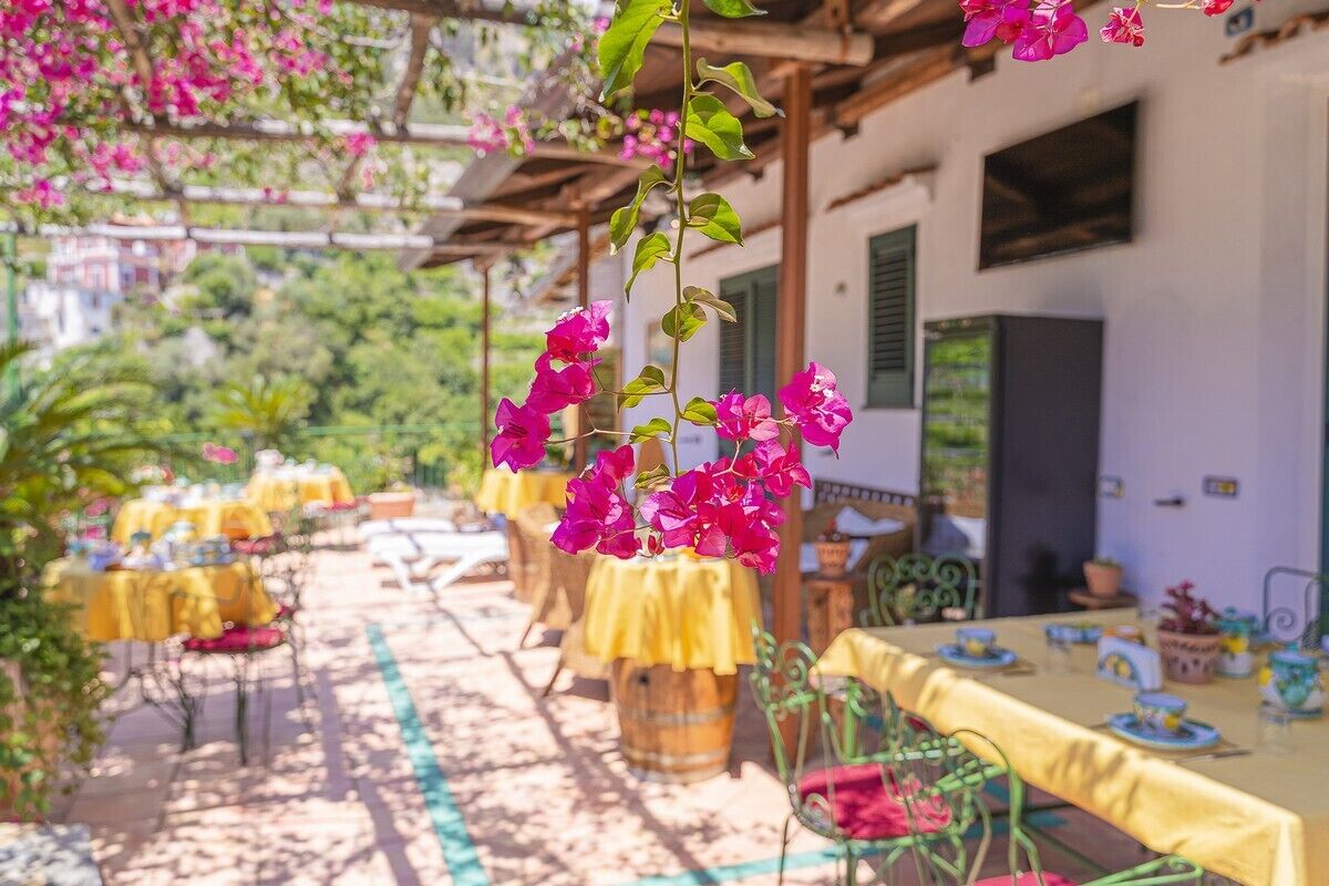 An outdoor dining area with yellow tables and chairs, surrounded by greenery and pink flowers.