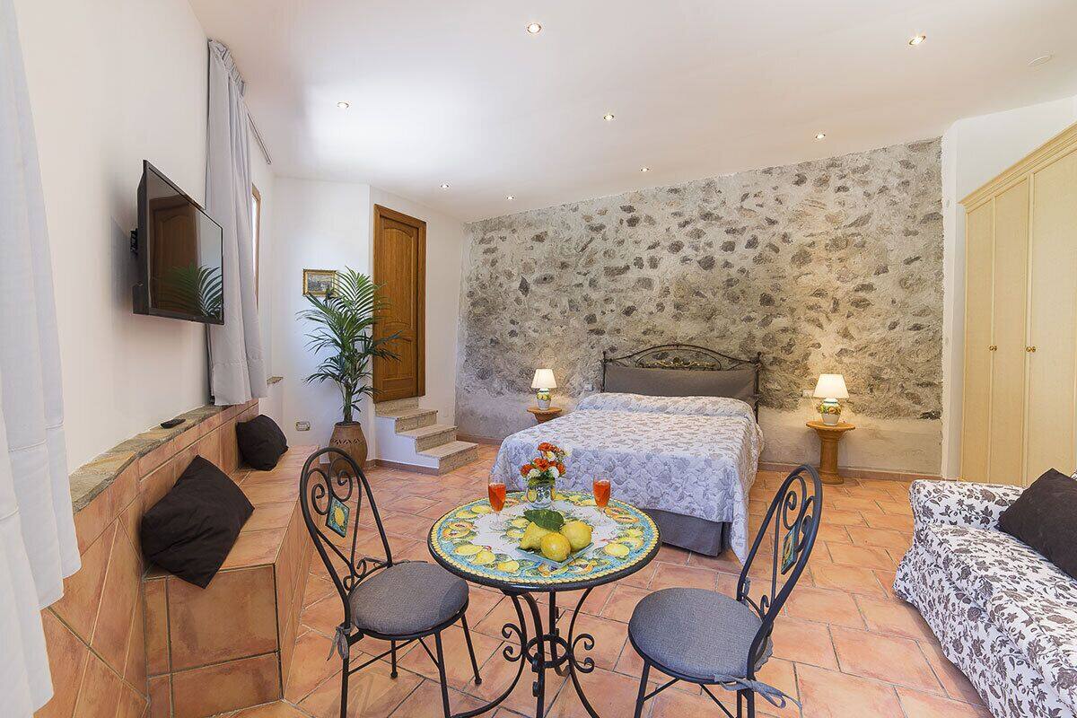 A bedroom with a stone wall, a bed with two bedside tables, a round table with chairs, and a sofa.