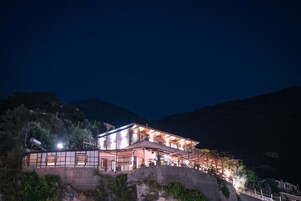 Un edificio de dos pisos con un balcón, situado en una ladera con montañas al fondo.