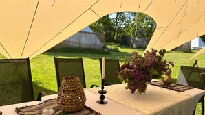 An outdoor dining setup with a table covered by a white tablecloth, surrounded by chairs with mesh backs. A woven basket and a vase with flowers are placed on the table. - Glamping in Stunning Bell Tent in Bohemia (Čmelíny)