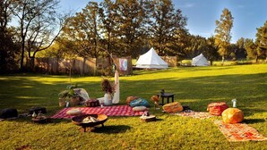 A grassy outdoor area with picnic setups, including blankets, cushions, and a fire pit. - Glamping in Stunning Bell Tent in Bohemia (Čmelíny)