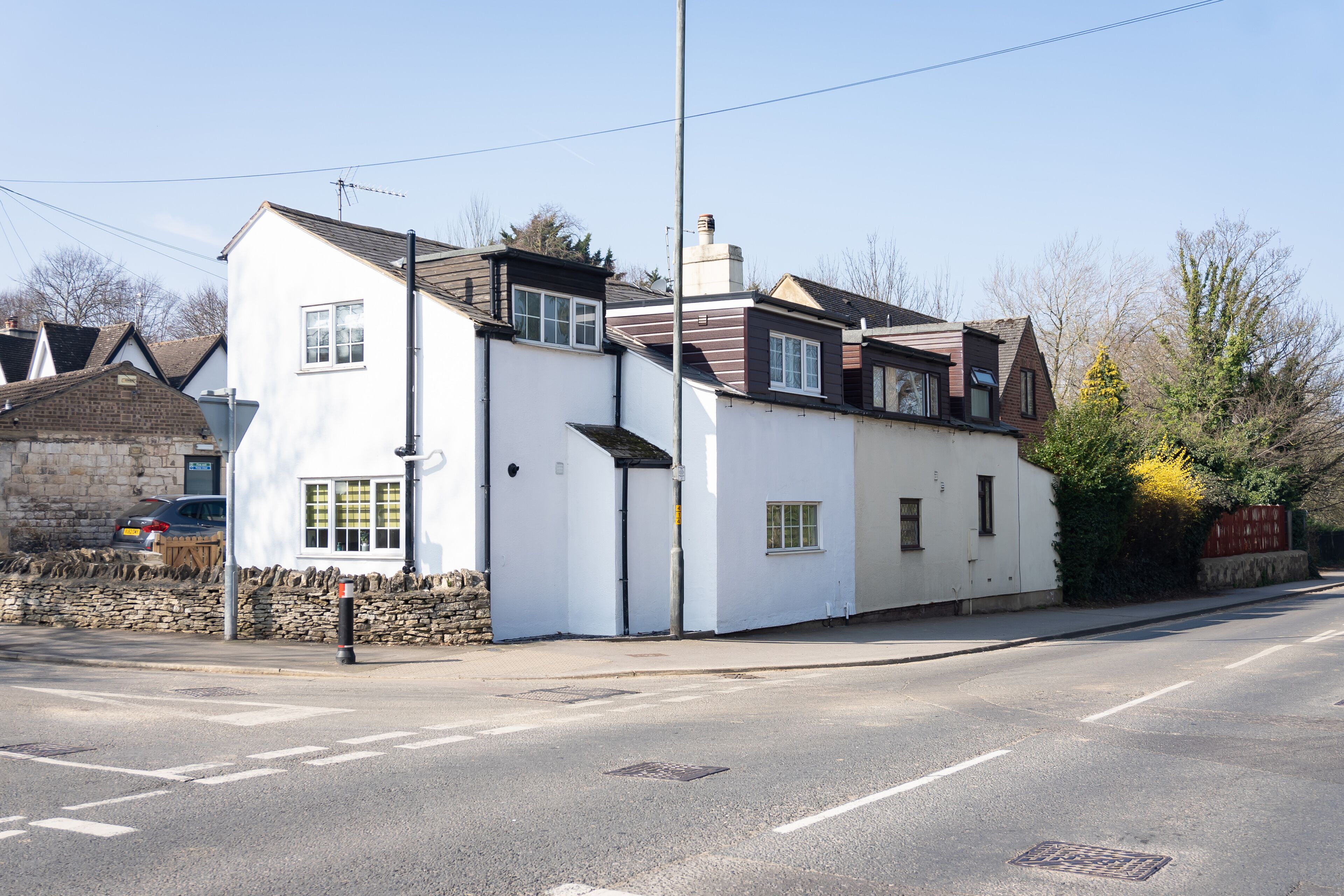 A two-story white building with a sloped roof, a stone wall, and a small garden area with a yellow flowering plant.