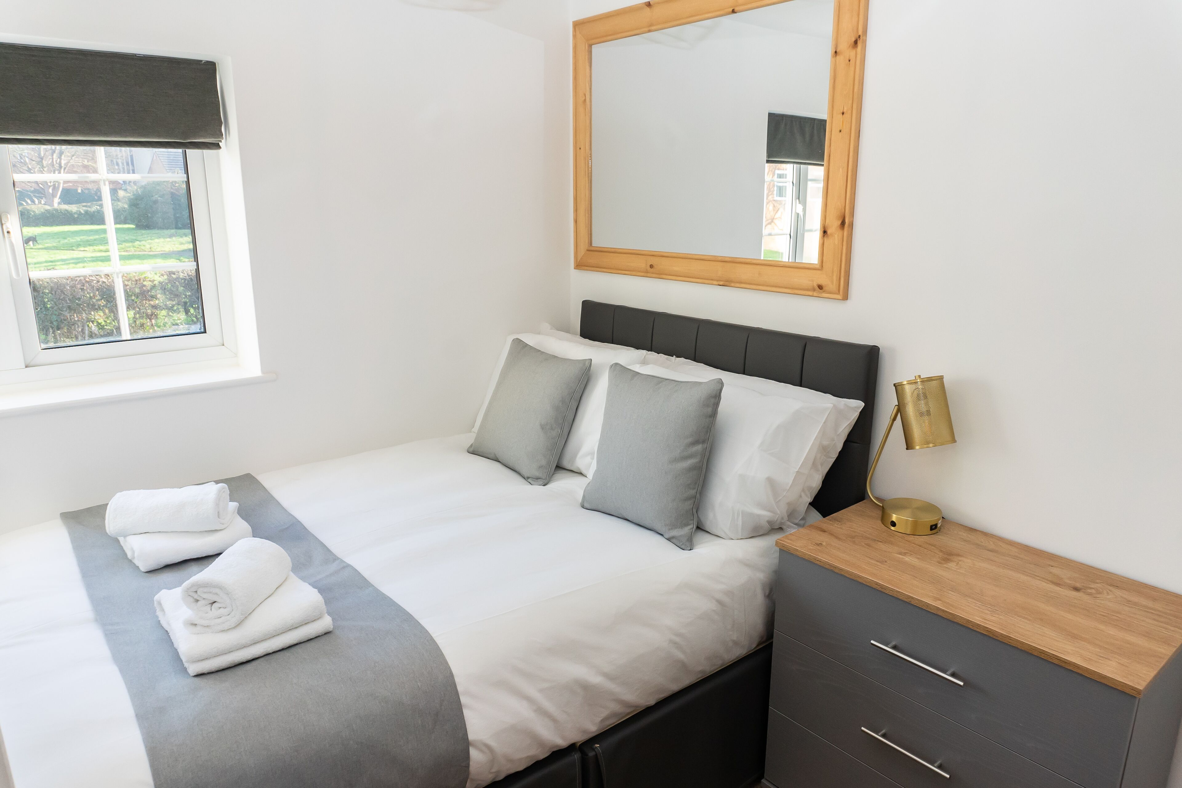 A neatly made bed with pillows, a bedside lamp, and a window with a view of greenery.