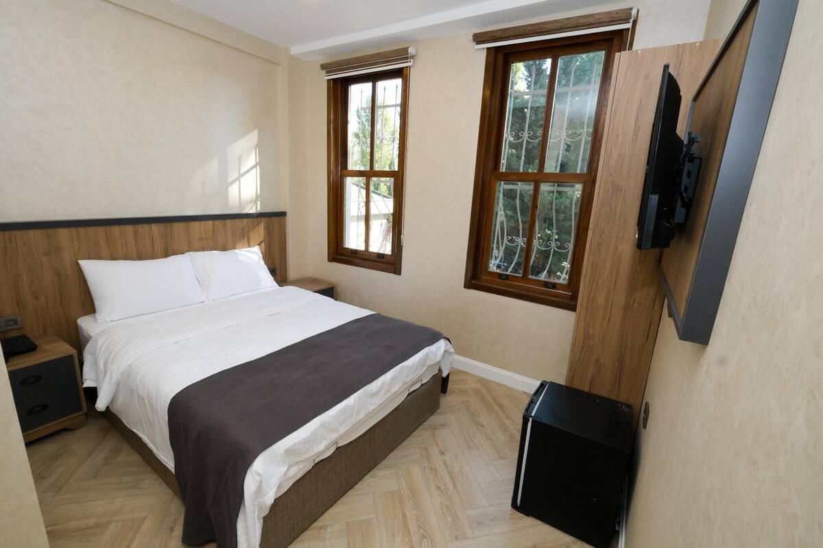 A bedroom with a bed, nightstand, TV, and two windows.