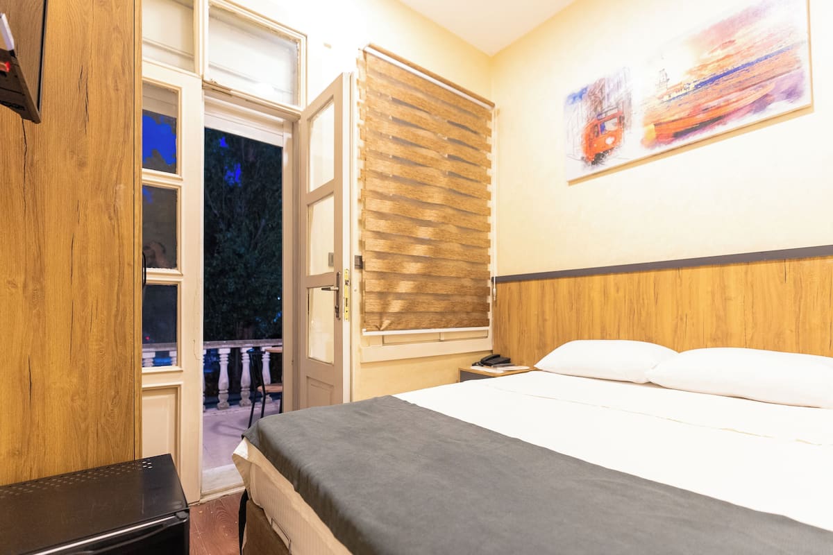 A hotel room with a bed, a wooden headboard, a window with blinds, and a painting on the wall.