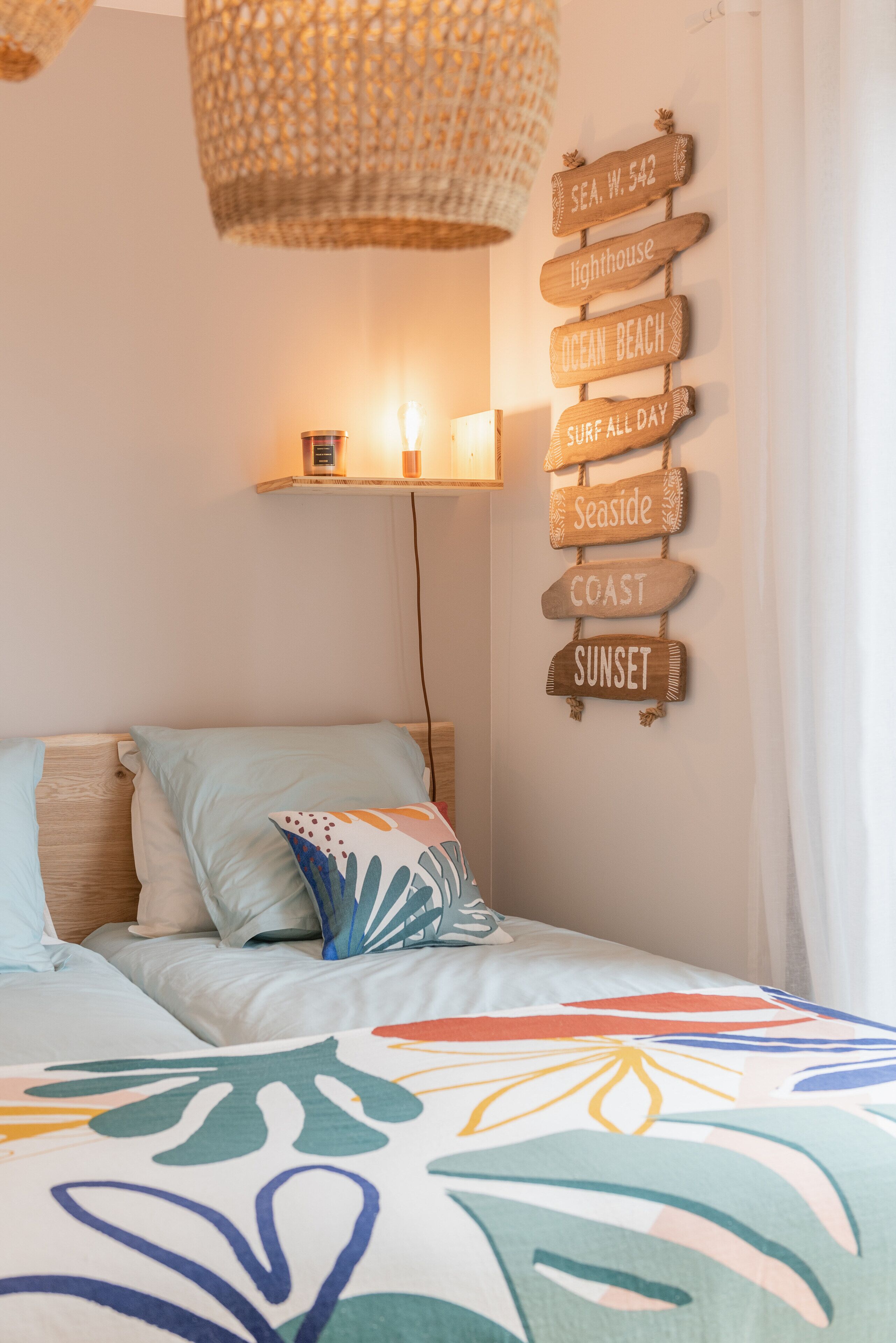Un dormitorio con cama, almohadas, una manta colorida, un cartel en la pared con destinos costeros y una lámpara colgante.