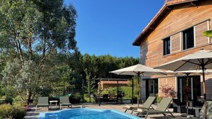 Une piscine entourée d’arbres et d’un bâtiment en bois, avec des chaises longues et des parasols à proximité.