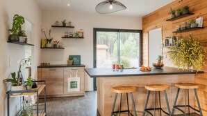 Une cuisine moderne avec des meubles en bois, un îlot central doté de tabourets, ainsi que diverses plantes et ustensiles de cuisine disposés sur des étagères.