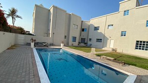 A swimming pool with a tiled deck, a covered area with a bench, and a building with multiple windows and a flat roof. - Villa 126 (Dubai)