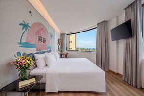 A hotel room with a large bed, a flat-screen TV, a desk with a laptop, and a view of the sea.