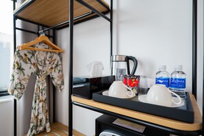 Ein Hotelzimmer mit einem Bett, einem Holzregal mit einem tropisch gestalteten Kleidungsstück, einer Kaffeemaschine, zwei Daquawa Wasserflaschen und einem Tablett mit Tassen und einem Glas.
