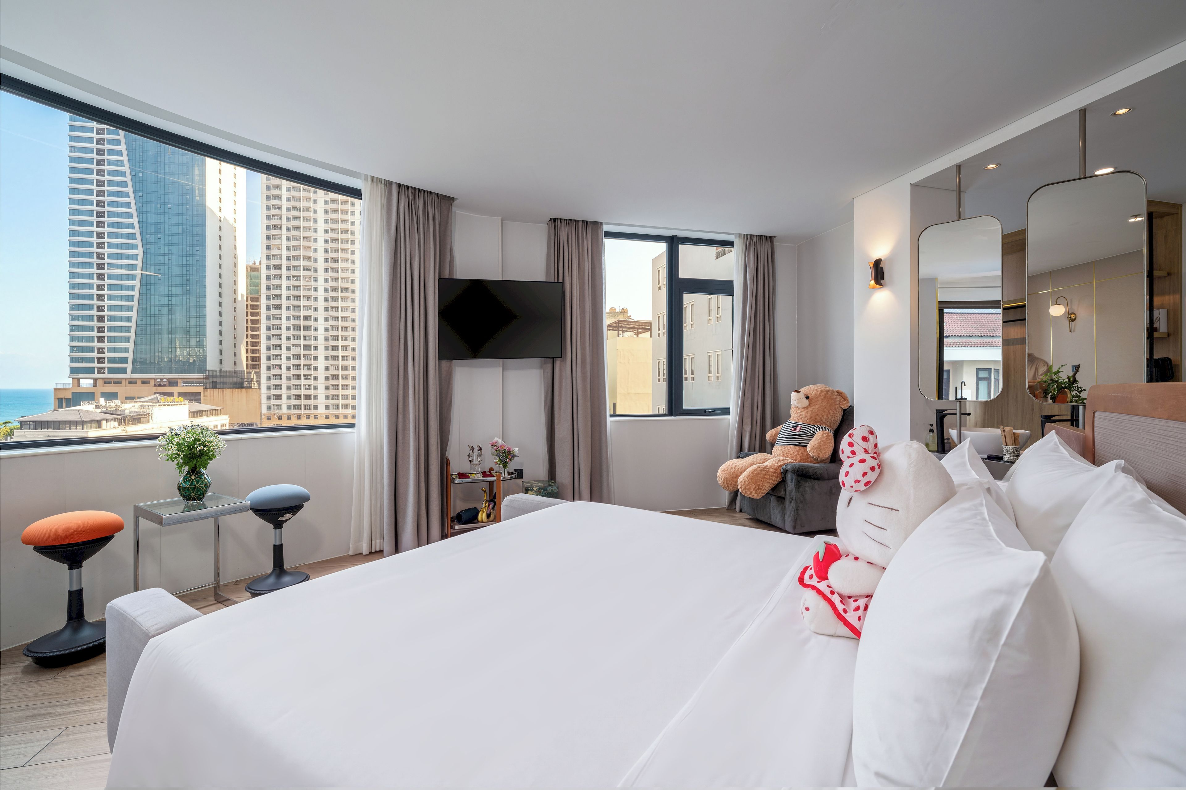 Une chambre d’hôtel avec un grand lit, un téléviseur à écran plat, une vue sur la ville et un balcon équipé d’une chaise et d’un ours en peluche.