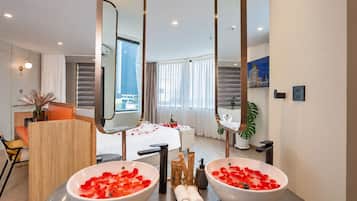 A hotel room with two large white basins filled with rose petals, a wooden desk with a vase, a bed with rose petals, and a large mirror.