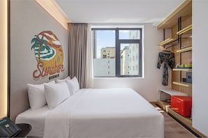 Une chambre d’hôtel équipée d’un lit, d’un téléphone, d’une fenêtre donnant sur des immeubles et d’un mur orné d’une œuvre d’art colorée.