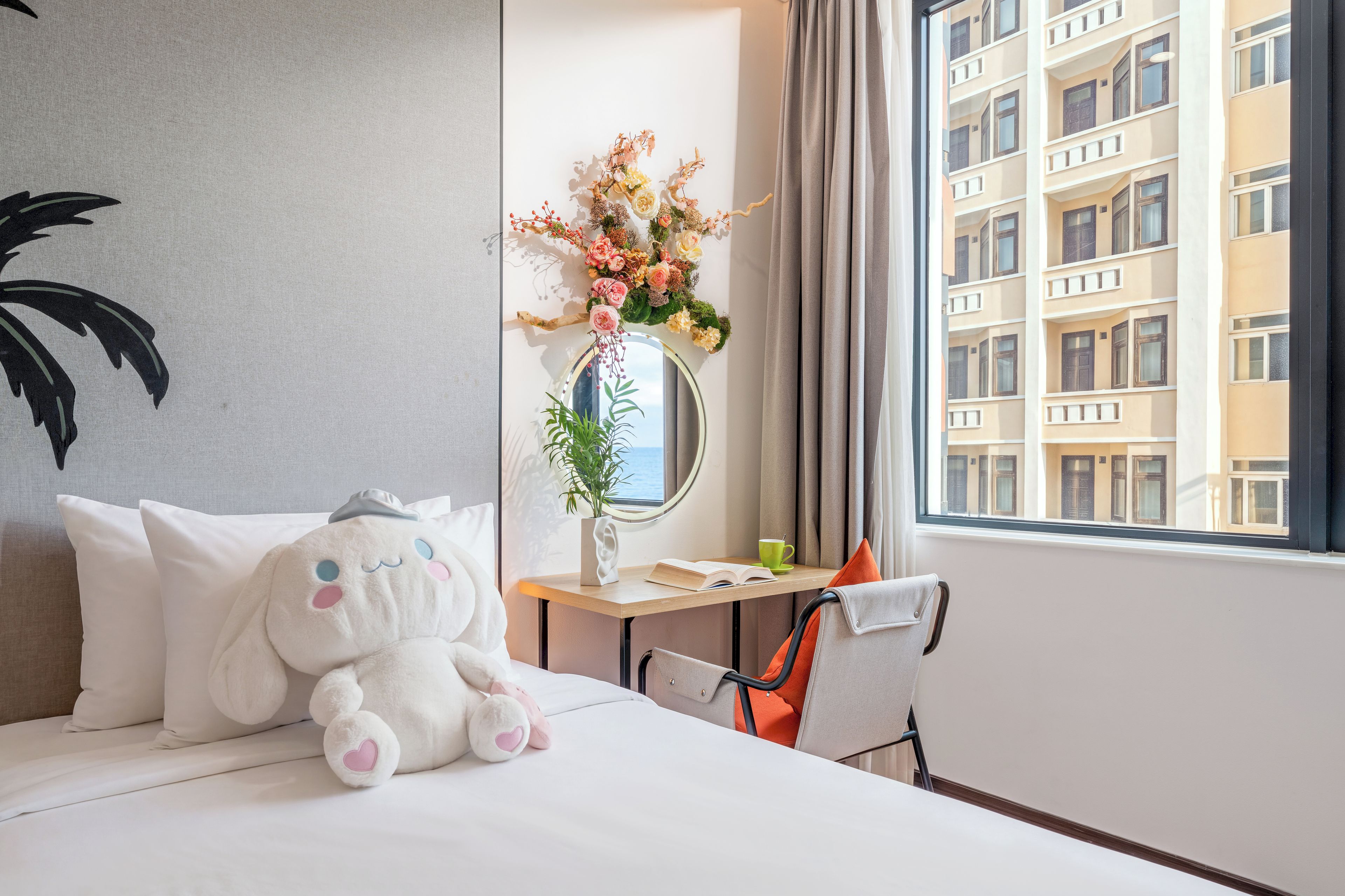 Habitación de hotel con cama, un peluche, un espejo, una mesita con una taza y un libro, y una ventana con vistas a edificios.