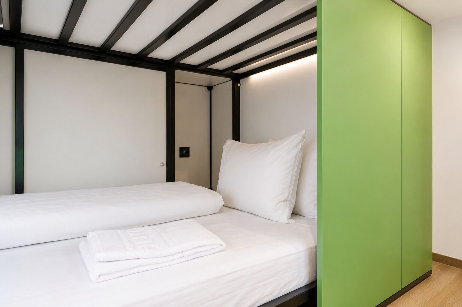Una cama individual con ropa de cama blanca y una toalla blanca doblada, un armario verde y suelo de madera.