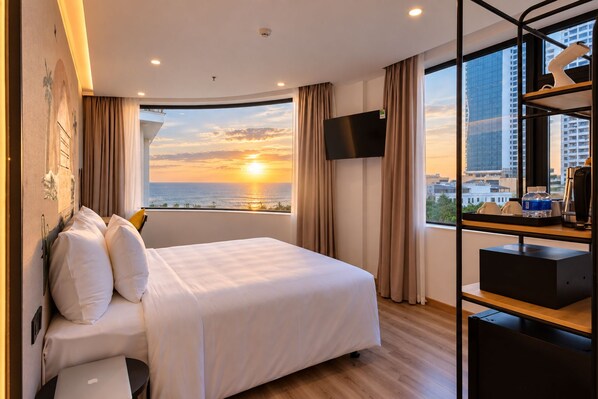 Ein Hotelzimmer mit Bett, Fernseher, Schreibtisch und Blick auf den Sonnenuntergang über dem Meer.