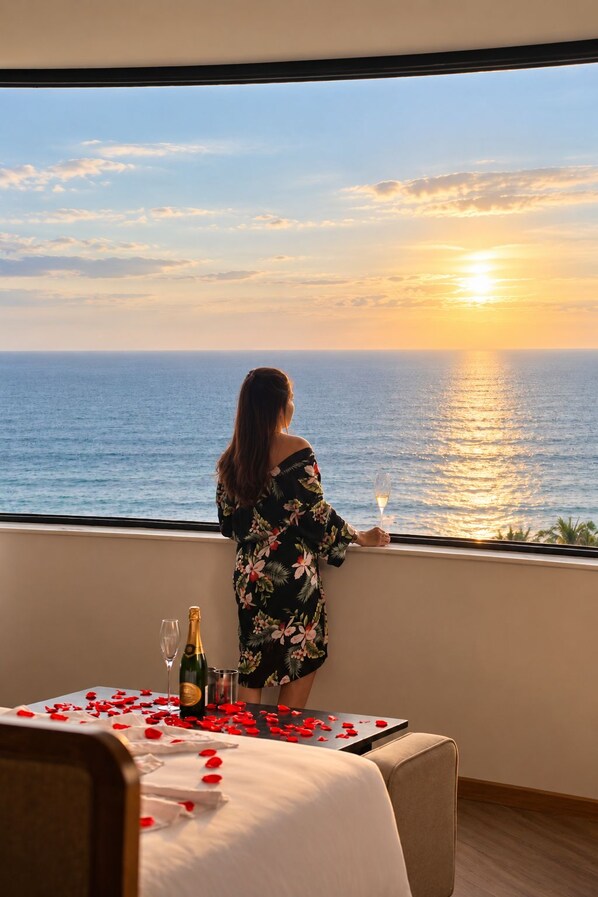A woman standing by a window overlooking the sea during sunset, with a table set with rose petals and champagne.
