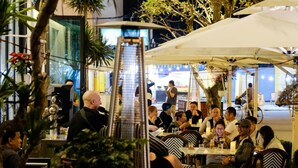 Un área de comedor al aire libre con mesas y sillas, rodeada de plantas y sombrillas.