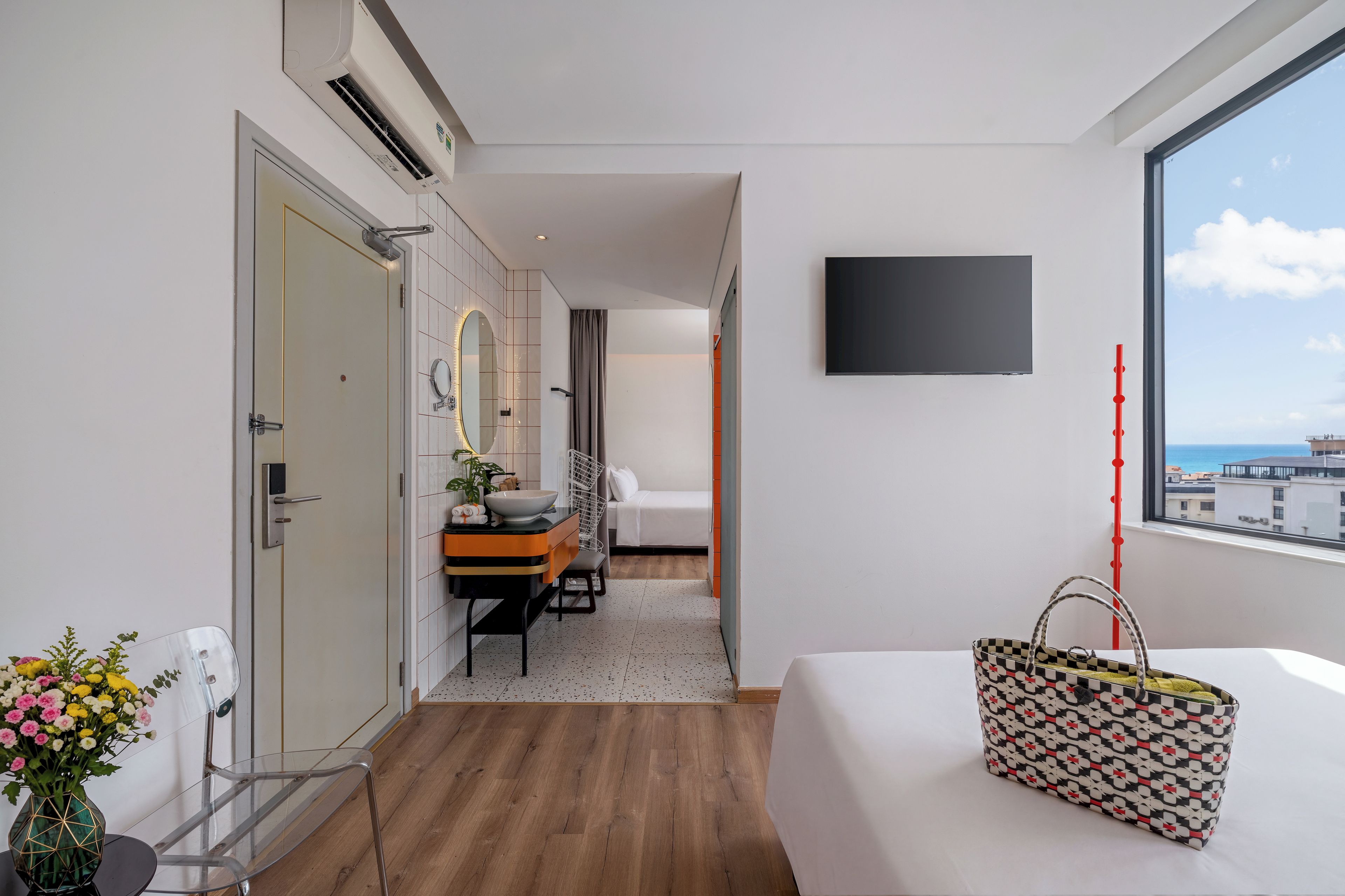 A modern hotel room with a bed, a bathroom with a mirror and sink, a small table with a plant, and a view of the city through the window.