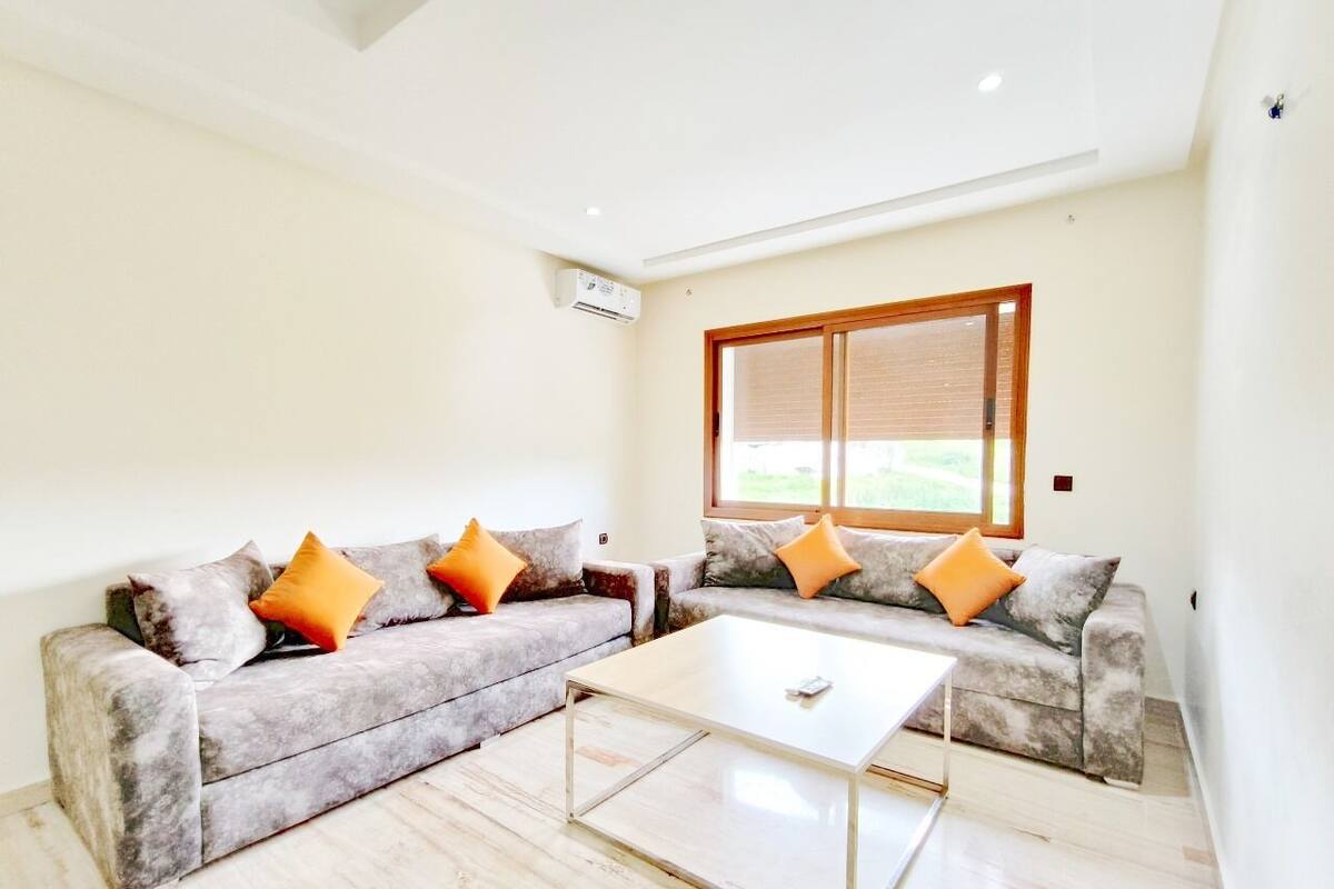 Un salon moderne avec deux canapés gris, chacun agrémenté de coussins orange, une table basse blanche et une grande fenêtre avec des stores.