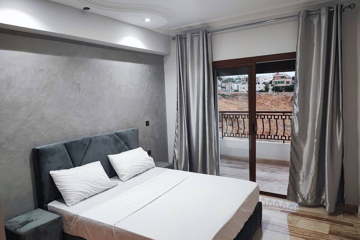 Une chambre à coucher avec un lit, une table de chevet, une fenêtre avec des rideaux et une vue sur un bâtiment et un paysage.