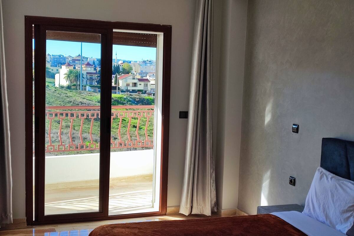 Une chambre à coucher avec un lit, une fenêtre donnant sur un balcon et des immeubles.