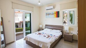 A bedroom with a bed, a nightstand, a lamp, a painting, and a window. - Villa Mesogeios Me16 (Paralimni)