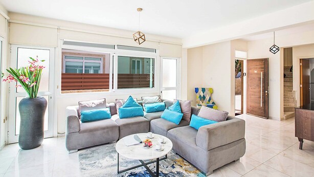 A spacious living room with a grey sectional sofa, a coffee table, and a large window with blinds. - Villa Mesogeios Me16 (Paralimni)
