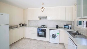 A compact kitchen with white cabinets, a built-in oven, a microwave, a washing machine, and a sink. - Villa Mesogeios Me16 (Paralimni)