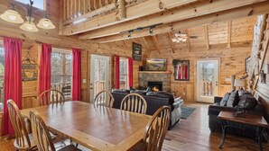 A rustic cabin interior with a wooden dining table, chairs, a fireplace, and a cozy seating area.