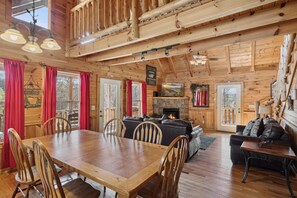 A rustic cabin interior with a wooden dining table, chairs, a fireplace, and a cozy seating area.
