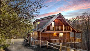 A wooden cabin with a balcony, surrounded by trees and a dirt path.