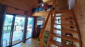 Une cabane en bois avec une chambre à coucher située en mezzanine, un escalier et un balcon agrémenté de mobilier d’extérieur.