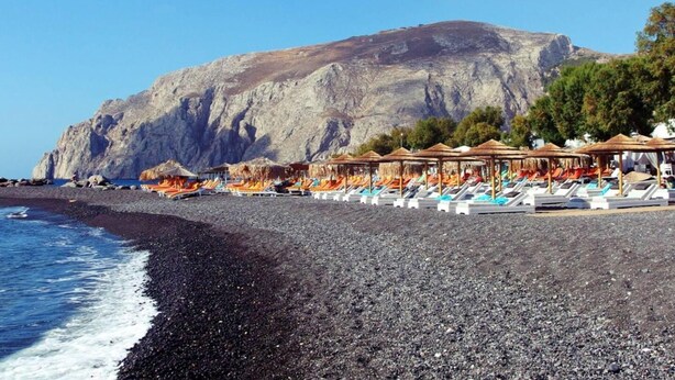 Ein Strand mit Sonnenschirmen und Liegestühlen, eine felsige Küste und ein Berg im Hintergrund.