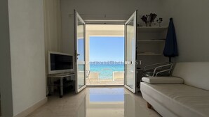 A modern living room with a white sofa, a TV, and a glass door leading to a balcony with a view of the sea. - Seafront Ground Floor (Zadar)