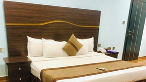 A neatly made bed with a wooden headboard, a bedside table with a phone, and a dark-colored wardrobe. - Ughelli Wetland Hotel (Ughelli)