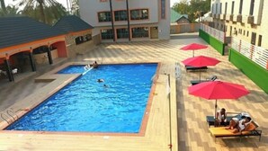 A rectangular pool with a wooden deck surrounding it, two red umbrellas, and a building with multiple windows in the background. - Ughelli Wetland Hotel (Ughelli)