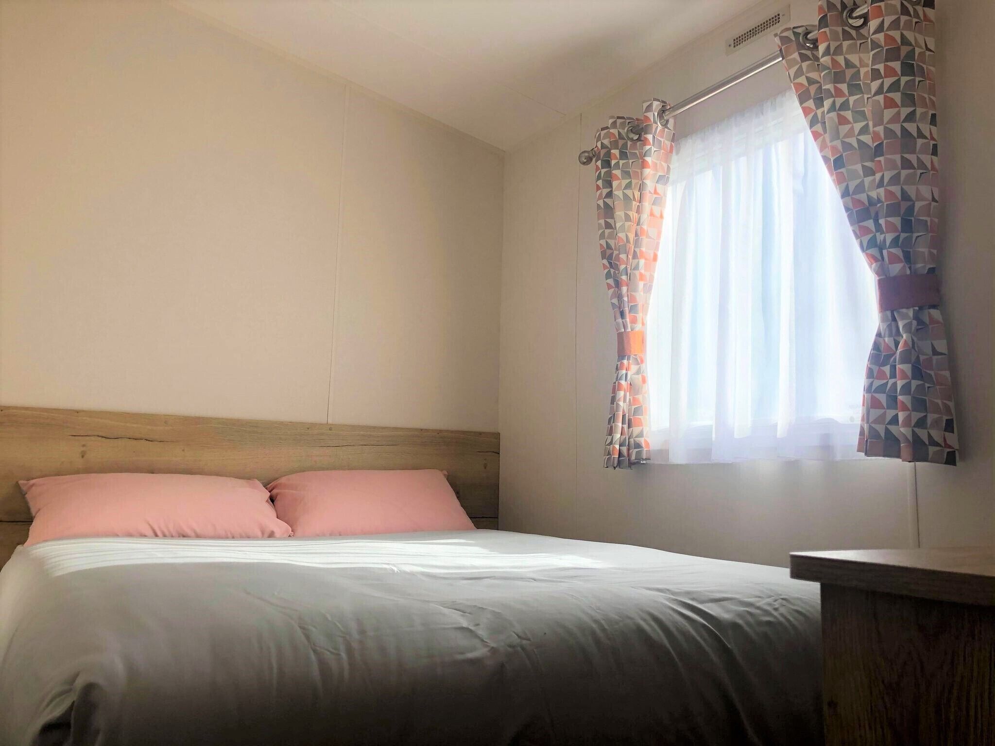 Une chambre à coucher avec une tête de lit en bois, un lit avec deux oreillers roses et une fenêtre ornée de rideaux à motifs.