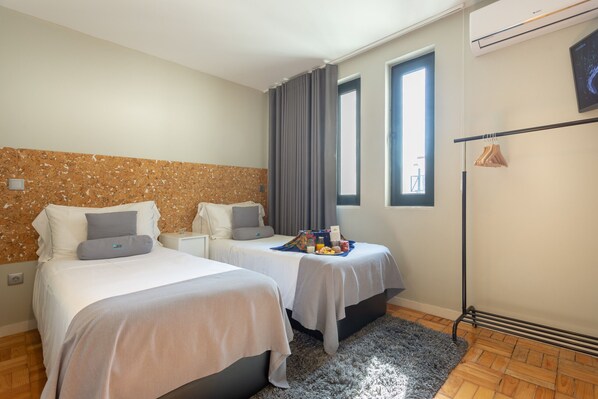 A hotel room with two beds, a grey rug, a wooden floor, a window with curtains, and a wall-mounted air conditioner.