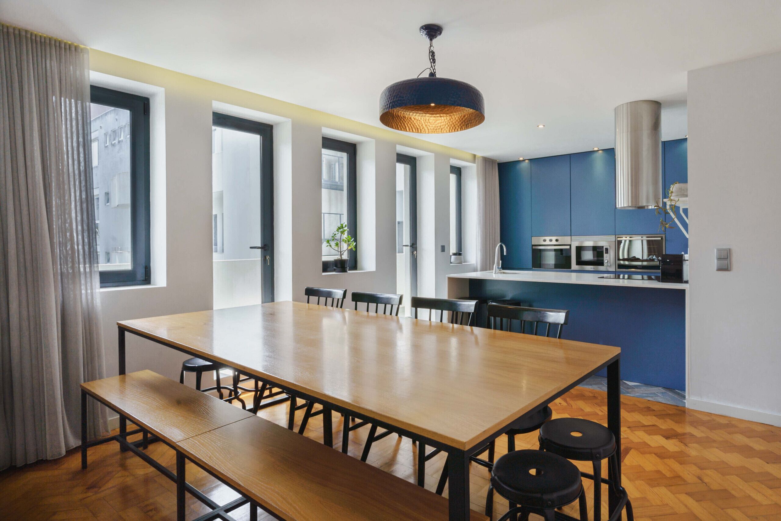 A modern kitchen with a central island, large windows, and a dining area with a wooden table and benches.