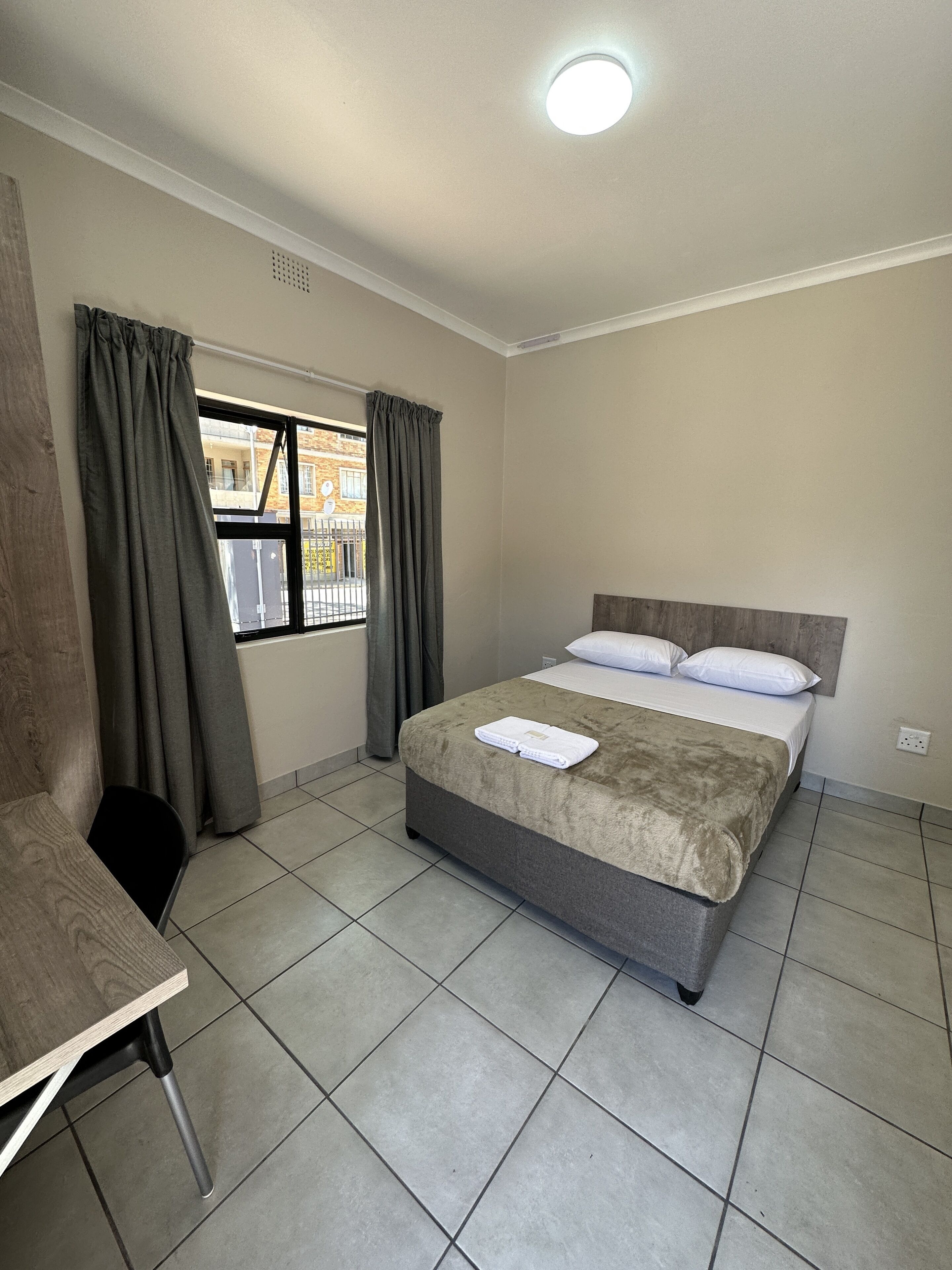 A hotel room with a bed, a chair, a window with curtains, and a view of buildings outside.