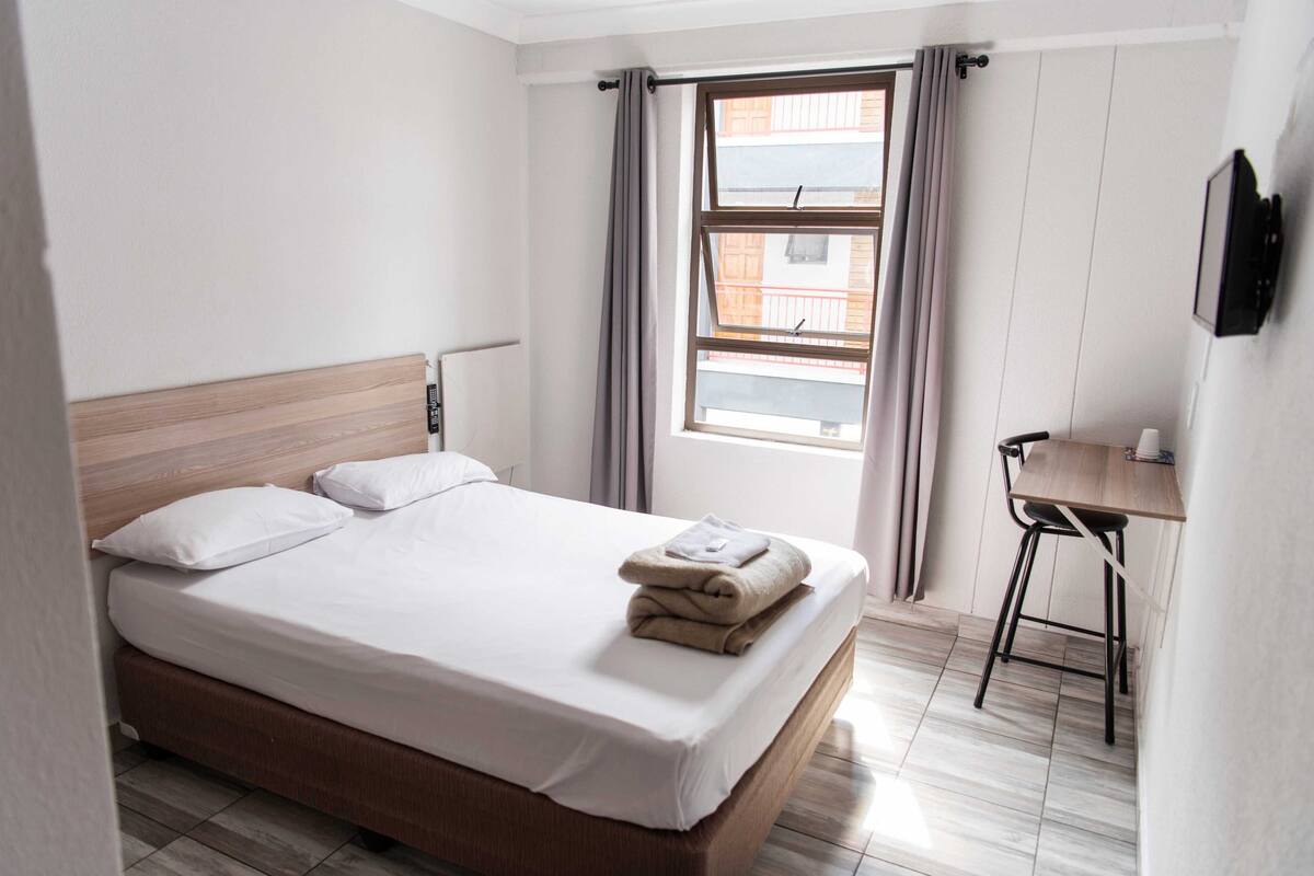 A neatly made bed with a wooden headboard, a small table with a chair, and a window with curtains.