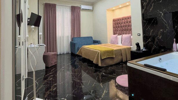 A modern hotel room with a large bed, a sitting area, a TV, and a bathroom with a marble floor. - My Luxury (Naples)