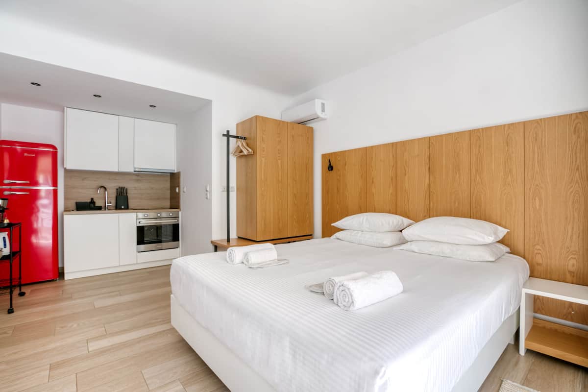 Une chambre d’hôtel moderne avec un grand lit, une tête de lit en bois, un réfrigérateur rouge, un coin cuisine et un plancher en bois.