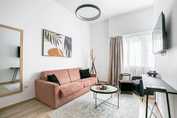 A modern living room with a pink sofa, a round coffee table, a flat-screen TV mounted on the wall, and a large window with sheer curtains. - Elia Kolonaki Luxury Apartments  (Athens)